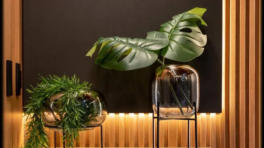 Nicho iluminado de um projeto do escritório, com vasos de vidro sobre bases metálicas, folhas de costela-de-adão e fundo ripado em madeira